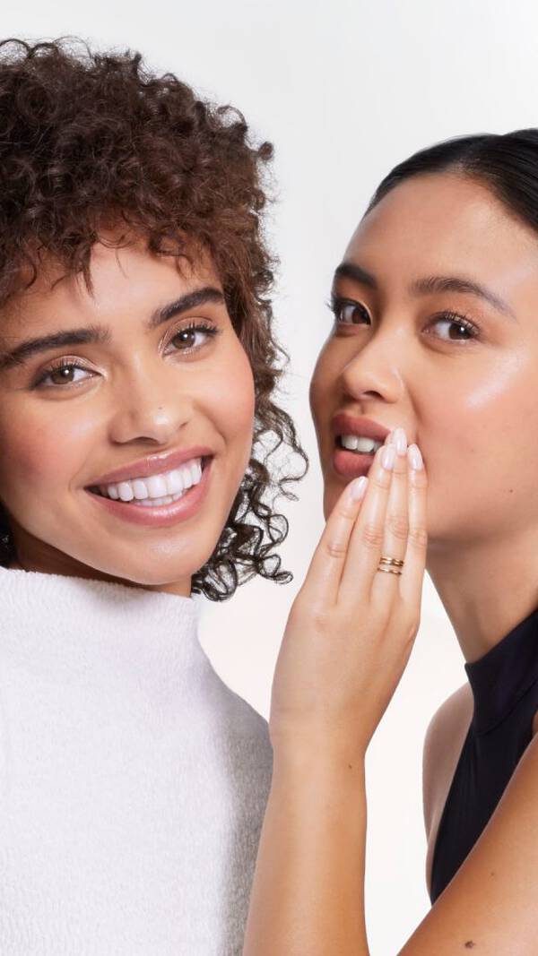 Deux femmes souriantes posent ensemble. L'une a des cheveux boucles et porte un haut blanc, tandis que l'autre a des cheveux lisses et un haut noir, chuchotant avec un sourire complice.