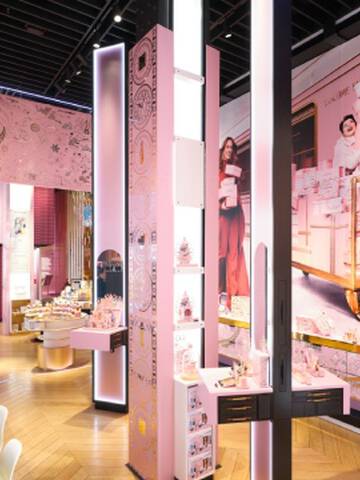 Vue interieure d'un magasin Lancome, avec des presentoirs roses et dores, des etageres de produits et une grande fresque murale representant des personnes avec des boites cadeaux roses.