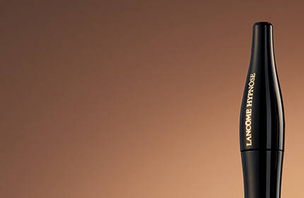 A close-up of three beauty products against a gradient brown background: a black eyeliner, a foundation bottle, and a red lipstick.