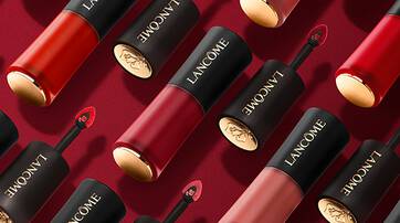 A collection of Lancome lip products in various shades of red and pink, featuring sleek black tubes with gold accents, arranged on a deep red background.
