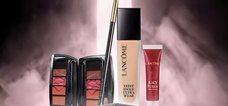 A display of three makeup products against a gradient brown background: a black eyeliner pencil, a beige foundation bottle, and a red lipstick tube, all branded with the Lancome logo.