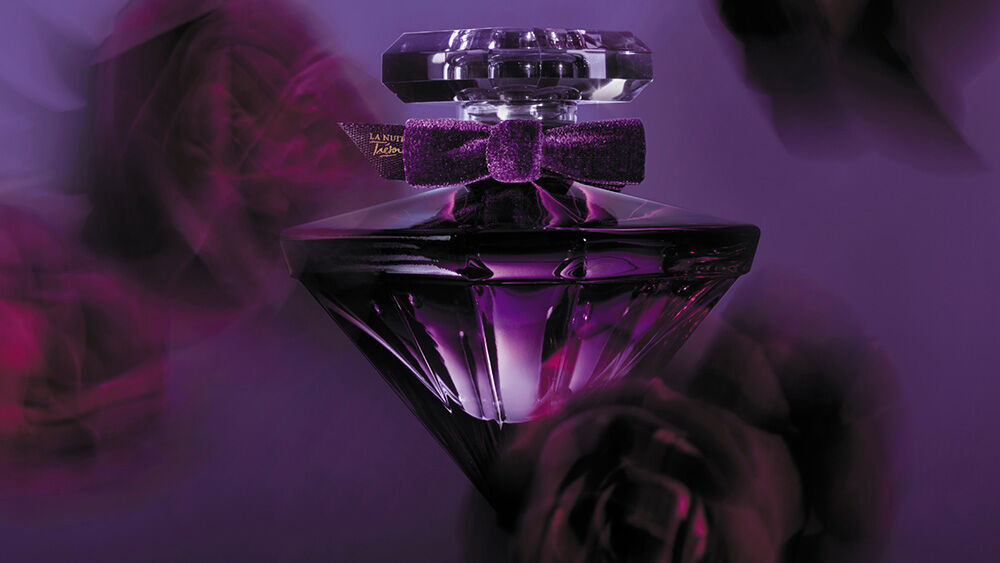 A stylish perfume bottle with a diamond shape, deep purple color, and a velvet bow, set against a blurred backdrop of purple roses.