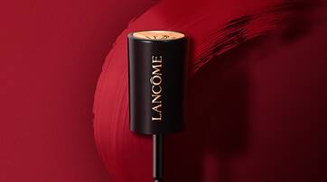 A lipstick tube from Lancome, featuring a sleek black design with a gold cap, is positioned above a vibrant red lip color smear on a matching background.