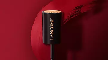 A lipstick tube from Lancome, featuring a sleek black design with a gold cap, is positioned above a vibrant red lip color smear on a matching background.
