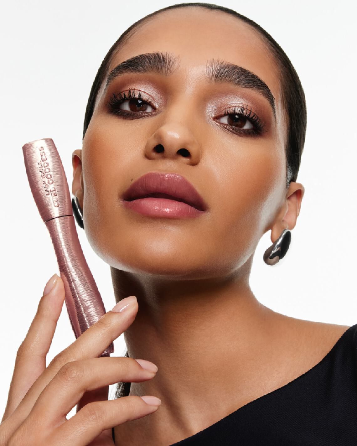 Femme tenant un tube de mascara Lash Idole Curl Goddess rose, mettant en valeur des cils volumineux et des levres vieux rose.