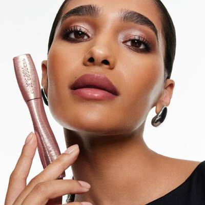 Femme tenant un tube de mascara Lash Idole Curl Goddess rose, mettant en valeur des cils volumineux et des levres vieux rose.