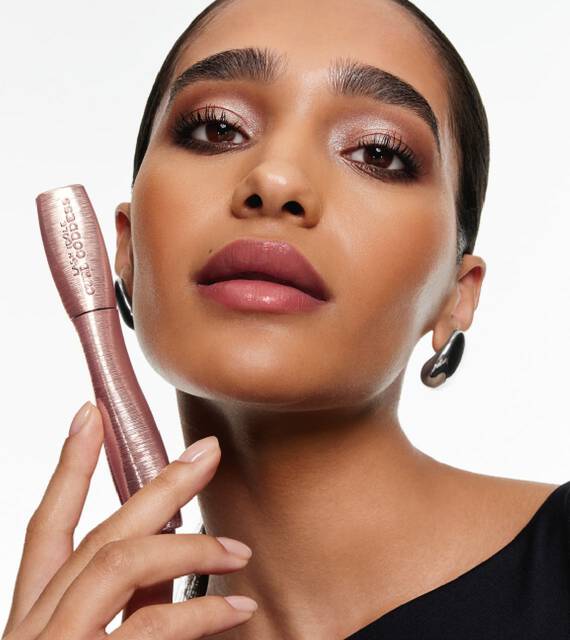 Femme aux cils volumineux et levres mauves tenant un tube de mascara rose.