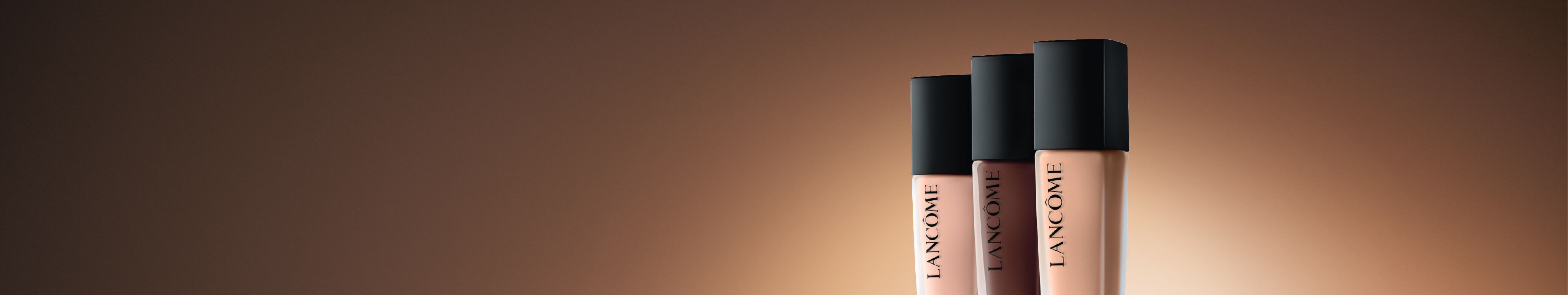 Three Lancome makeup products are displayed prominently against a warm, gradient background. The items include a compact with a blush, a bottle of foundation labeled "Teint Idole Ultra Wear," and a tube of concealer.