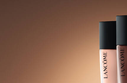 Three Lancome makeup products are displayed prominently against a warm, gradient background. The items include a compact with a blush, a bottle of foundation labeled "Teint Idole Ultra Wear," and a tube of concealer.