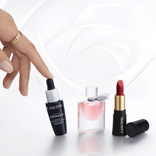 Une main touche un flacon de serum de beaute, entoure d'un parfum et d'un rouge a levres, le tout sur un fond blanc.