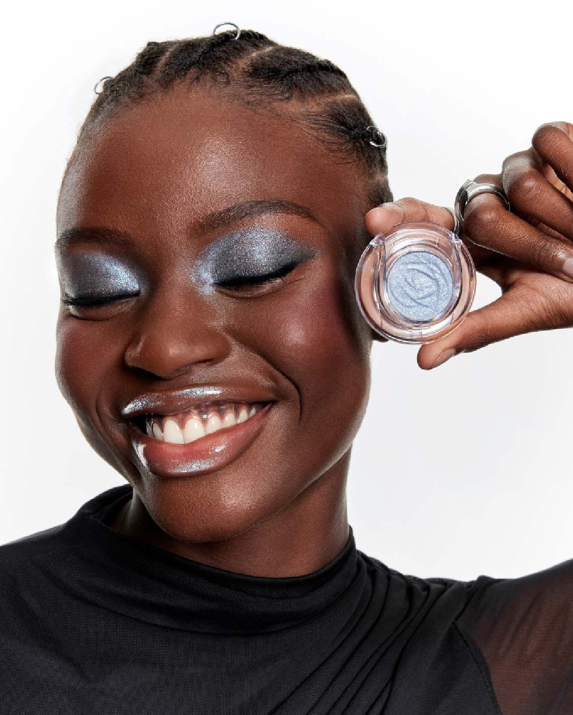 Femme souriante aux yeux clos, portant un fard a paupieres bleu scintillant et tenant un boitier d'ombre a paupieres assorti.