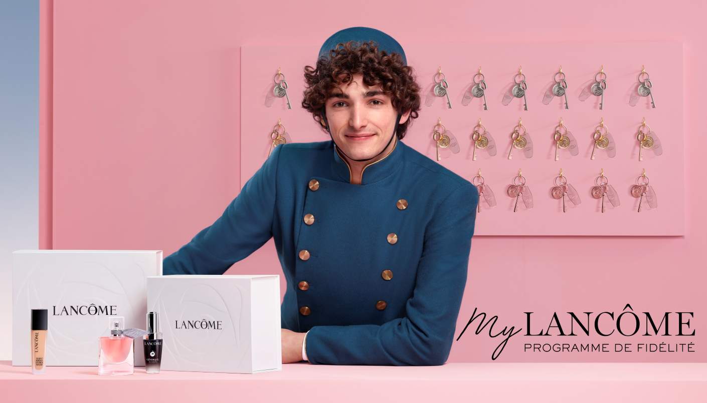 Une main tendue vers un flacon de parfum Lancôme et un mascara sur une boîte blanche ornée d'un ruban, mettant en valeur des produits de beauté de luxe.