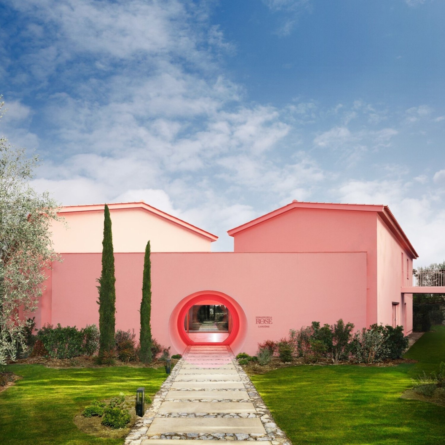 Maison moderne rose avec un grand cercle d'entrée, entourée de verdure et sous un ciel bleu et nuageux.