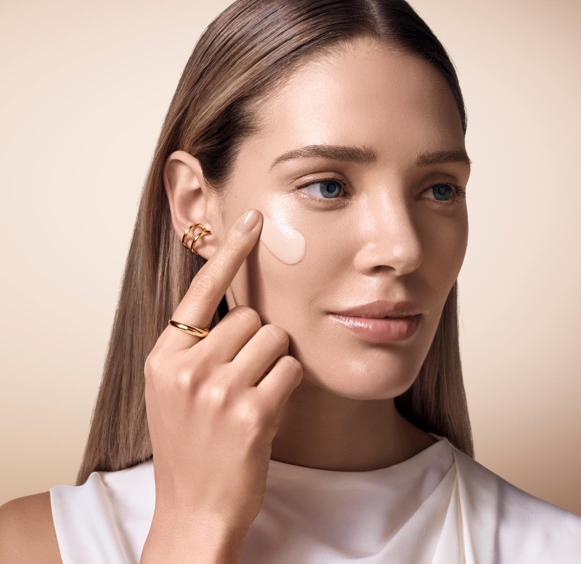 Une femme applique une crème de soin sur son visage, affichant une peau éclatante, avec des bijoux délicats.