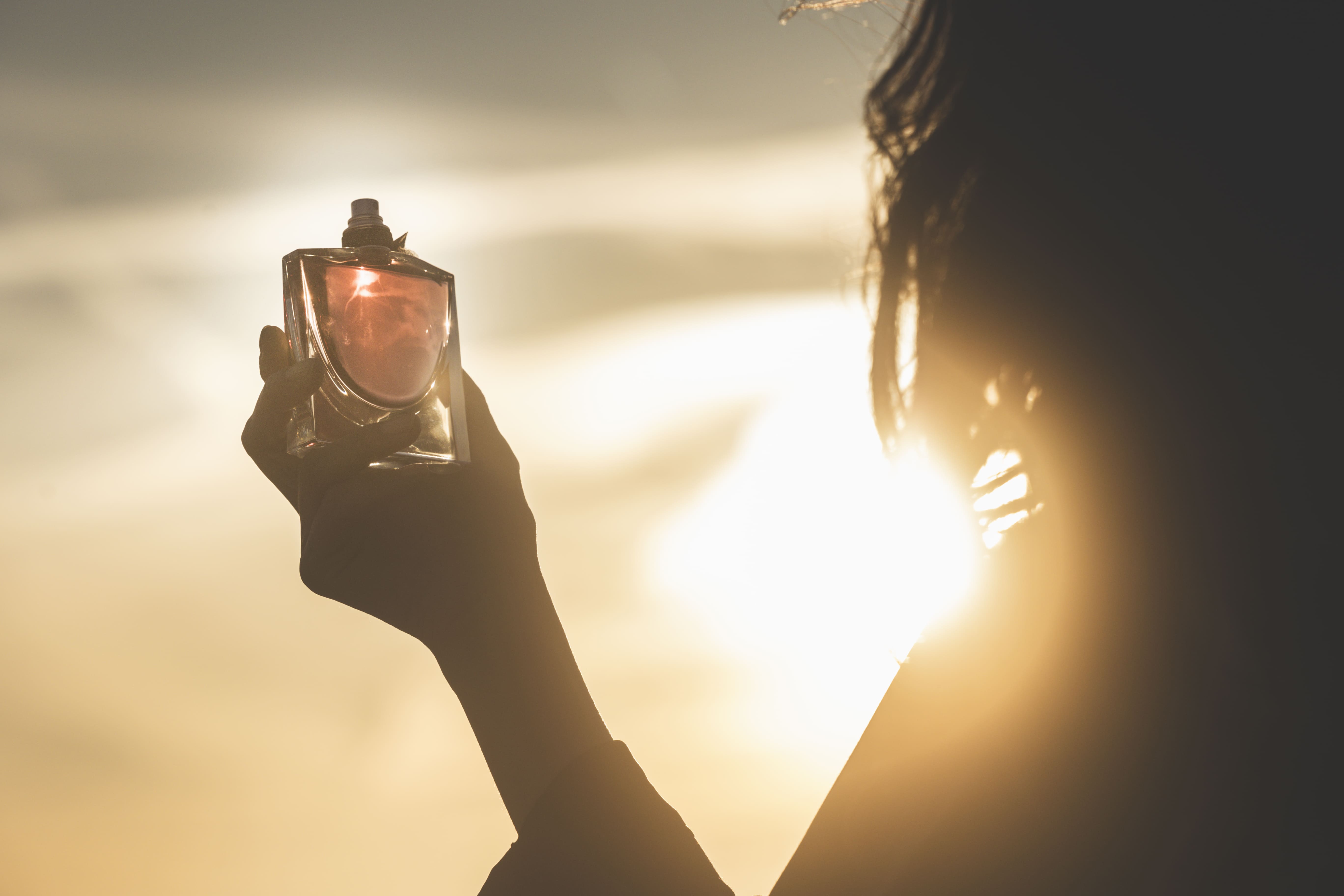 Une silhouette féminine tient un flacon de parfum, silhouettée contre un soleil couchant.