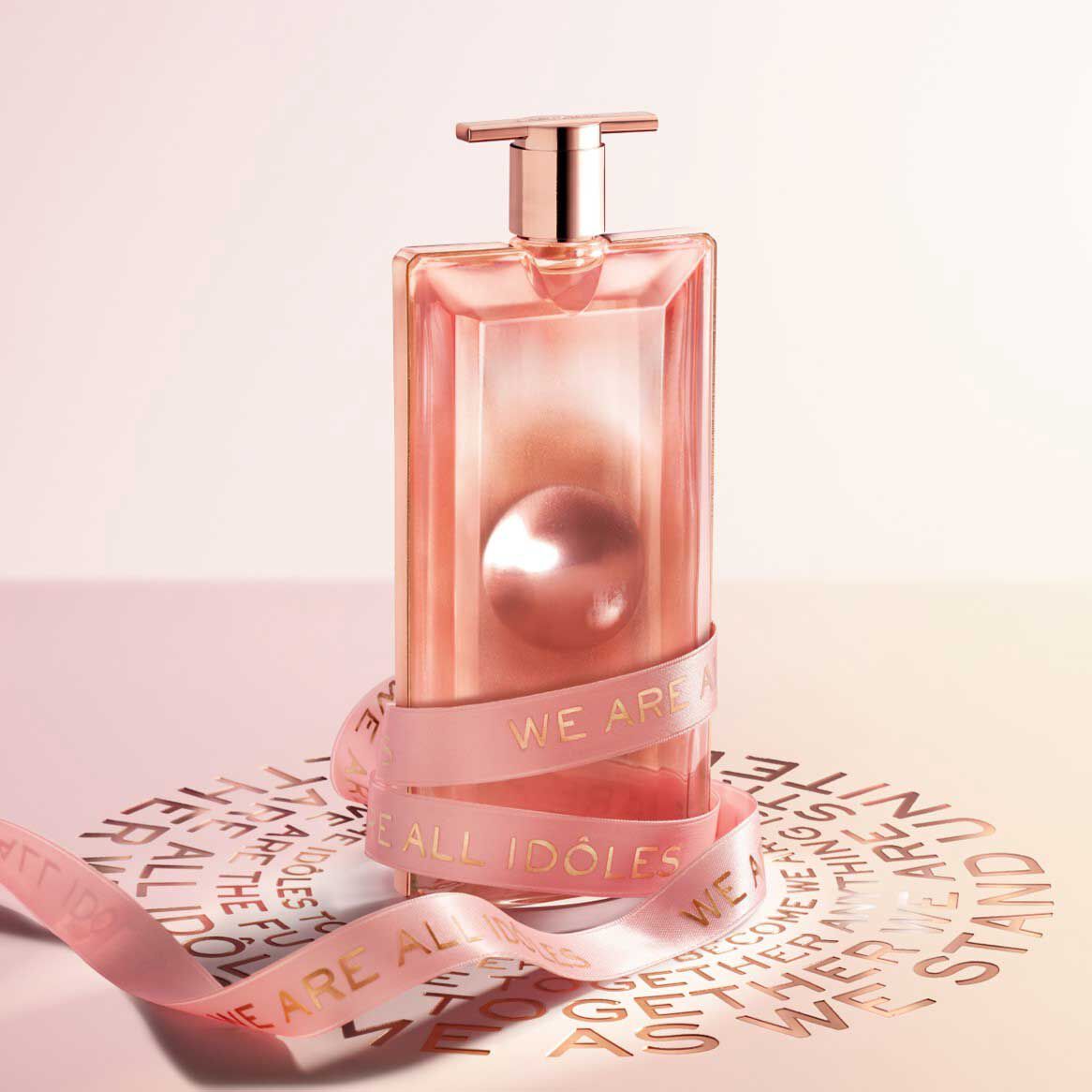 Flacon de parfum élégant en verre rose, avec un ruban portant l'inscription 'WE ARE ALL IDÔLES', sur un fond artistique.