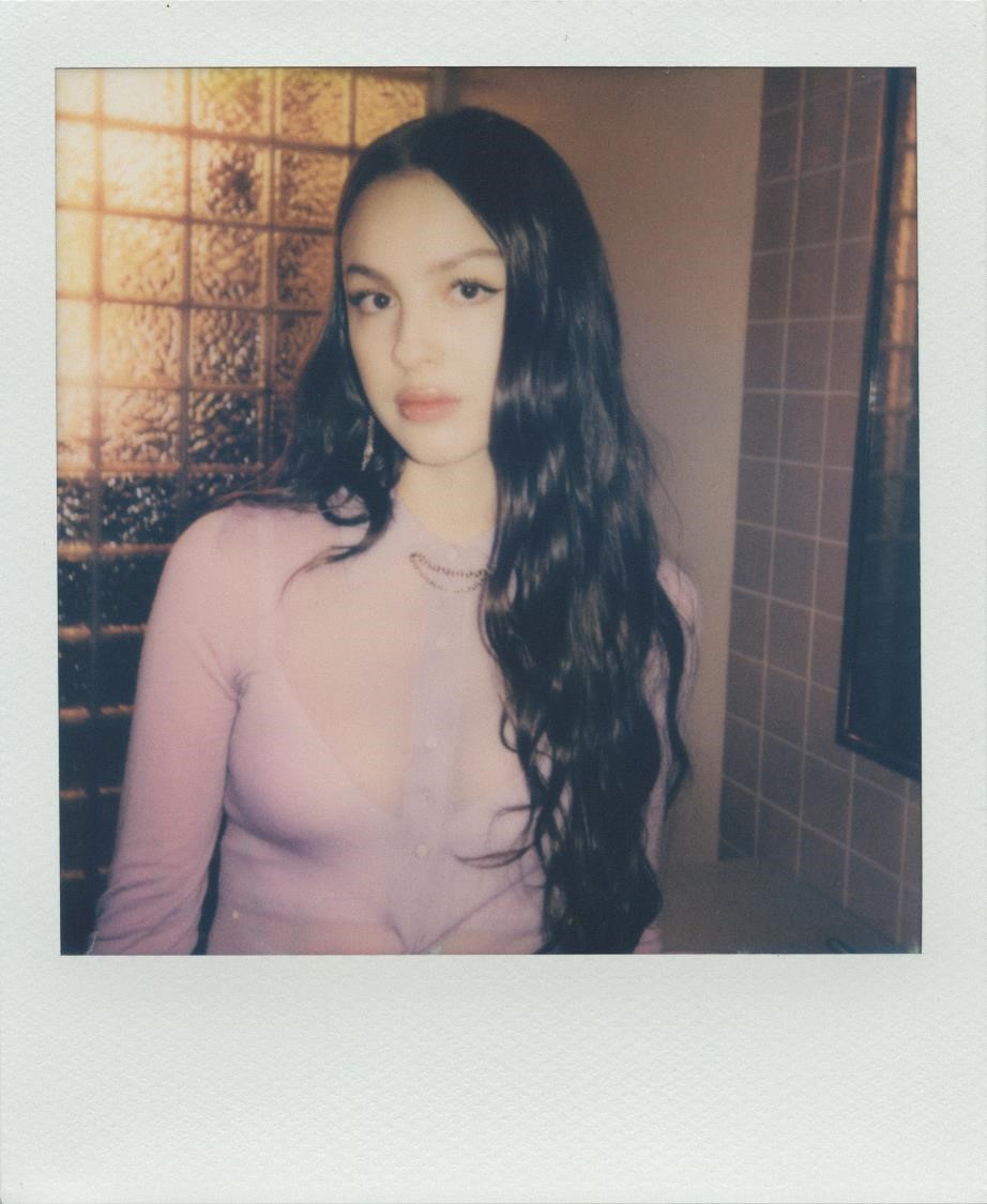 Portrait d'une jeune femme avec des cheveux longs et ondulés, portant un haut rose, devant un mur à carreaux.
